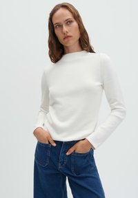 Frau mit welligem Haar, die ein weißes Langarmshirt und dunkelblaue Jeans trägt, die Hände lässig in den Vordertaschen, neutraler Hintergrund.