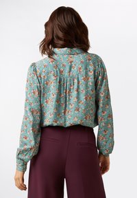 Bluse mit Blumenmuster aus türkisfarbigem Stoff, mit Knopfmanschette und gerafftem Rückenteil. Verfügt über kleine, mehrfarbige Blumenmuster auf der gesamten Fläche.