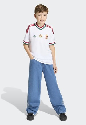 Garçon debout, une main dans la poche, portant un maillot de sport blanc avec des rayures rouges et vertes, un jean large bleu et des chaussures noires.