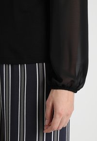 Svart topp med genomskinliga långa ärmar; ribbade muddar. Kombinerad med randiga byxor i marinblått, grått och vitt. Mjuk textur och lagerdesign.