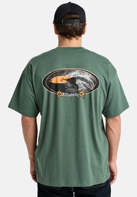 Green cotton t-shirt with short sleeves. Back features a graphic of a surfer in an orange and black oval. Billabong logo in floral detail.
