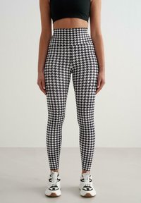 Legginsy z wysokim stanem w czarno-biały wzór pepitki, o dopasowanym kroju i gładkiej fakturze, zestawione z białymi i czarnymi sneakersami.