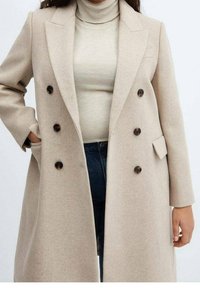 Beige uldblanded dobbeltbrystet frakke med store brune knapper, slidser i kraven og sidelommer. Båret over en lys turtleneck og jeans.