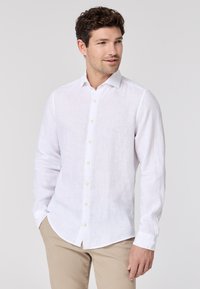 Weißes Leinen-Hemd mit langen Ärmeln, spitzem Kragen, durchgehender Knopfleiste, weichem Material und abgerundetem Saum, kombiniert mit beigen Hosen.