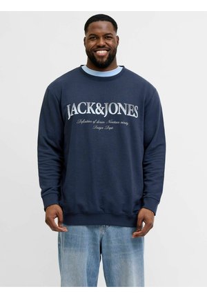 Uśmiechnięty mężczyzna w granatowej bluzie z napisem "JACK&JONES" i jasnoniebieskich dżinsach, stojący na jednolitym jasnym tle.