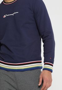 Mörkblå sweatshirt med ribbad krage och flerfärgade randiga manschetter. Har "Champion"-logotyp i vitt, tillverkad av en bomullsmix.