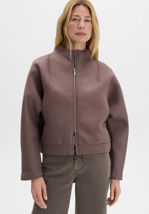 Frau trägt eine braune Jacke mit Reißverschluss und graue Hose und steht vor einem schlichten weißen Hintergrund.