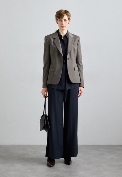 Frau in einem grauen Blazer, navy-blau gestreifter Bluse und weiter Hose, die eine schwarze Handtasche hält und vor einer hellgrauen Wand steht.