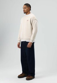 Beige sweatshirt met een ruime pasvorm, ronde hals en geborduurd logo, gecombineerd met donkerblauwe wijde jeans en bruine veterlaarzen.