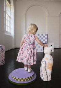 Vaikas rožinės gėlių suknelės stovi ant apvalaus purpurinio pjedestalo ir tiesia rankas link baltos popieriaus-macijo katės su oranžiniais akcentais.