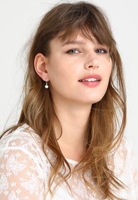 Model with long brown hair wearing delicate silver earrings with a small pearl. She has a light lace top and natural makeup.