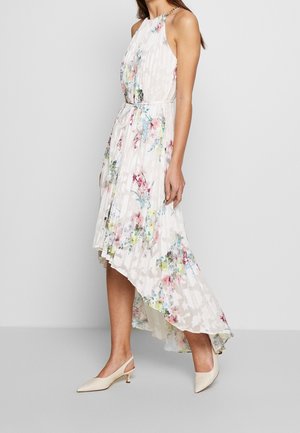 Mujer con vestido blanco sin mangas de flores con dobladillo alto-bajo y pliegues, y zapatos slingback beige de tacón bajo, de pie frente a un fondo liso.