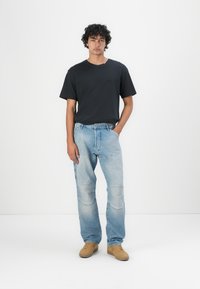 Zwarte T-shirt met korte mouwen, losse lichtblauwe denim jeans met vervaagde accenten en kniebeschermers, en beige sneakers. Effen achtergrond.