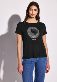 T-shirt noir à manches courtes avec un motif graphique circulaire blanc et le mot "SOLEIL" imprimé en dessous. Tissu simple et léger.