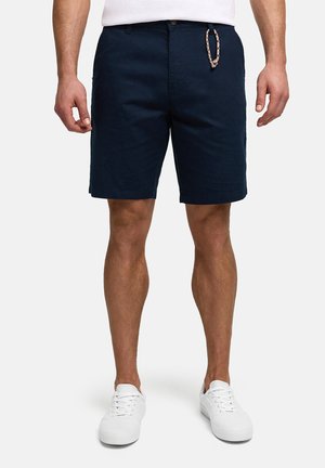Mann iført marineblå knelange shorts, hvite joggesko og en hvit skjorte med en flettet nøkkelring festet til shortsen.
