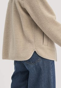 Beige Wolljacke mit einem abgerundeten Saum, strukturiertem Oberflächenmaterial und seitlichen Taschen, kombiniert mit dunkelblauen Denim-Jeans.