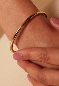 Gouden metalen bangle armband, slank ontwerp, gladde afwerking, gedragen om een pols, handen die voorzichtig vasthouden, lichte reflectie zichtbaar.