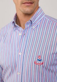 Camisa de algodón de botones con rayas verticales azules y rojas, un cuello con botones y un pequeño logo bordado en el lado izquierdo del pecho.