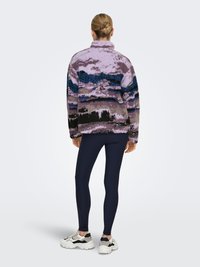 Paarse en zwarte fleece trui met een gestructureerd landschapspatroon; gecombineerd met marineblauwe leggings en stevige witte sneakers. Achteraanzicht.