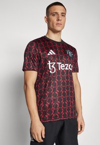 Manchester United -teemattu paita, musta punaisella geometrisella printillä, lyhythihainen, pyöreä kaula-aukko, jossa valkoinen logo ja teksti.