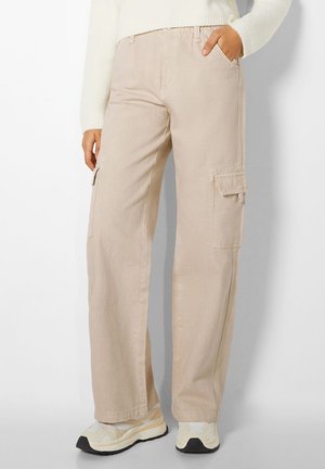 Person wearing beige wide-leg cargo pants with side pockets and white sneakers, hand in pocket against plain background.
