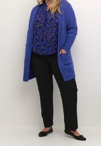 Langer, offener Cardigan aus strukturiertem blauen Strick, mit zwei aufgesetzten Taschen vorne, kombiniert mit einer blauen Bluse mit floralen Mustern und schwarzen Hosen, abgerundet mit schwarzen Ballerinas.