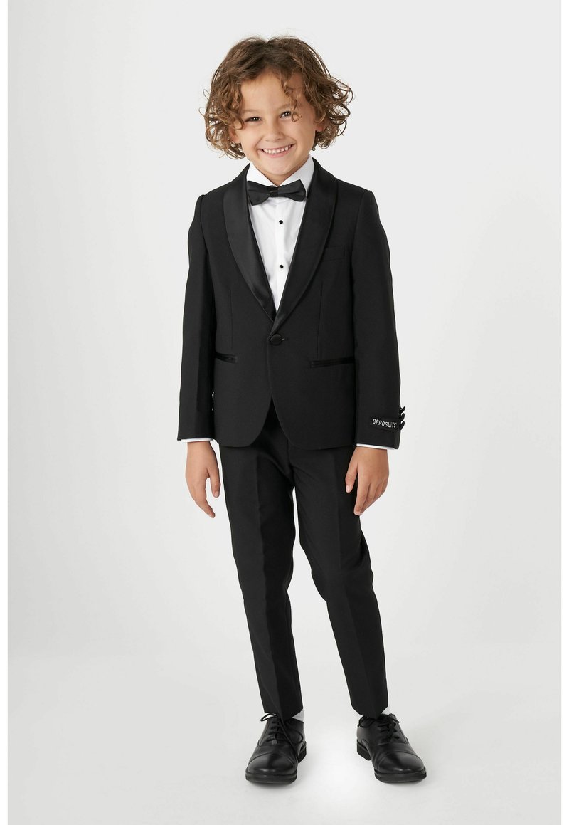 Tuxedo noir avec revers en satin, chemise blanche à boutons noirs et nœud papillon noir. Complété par des chaussures de soirée noires.