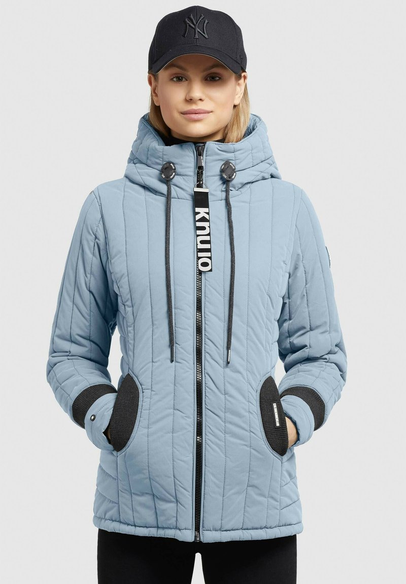 Helles blaues gesteppter Jacket mit hohem Kragen, schwarzen Akzenten an den Taschen und einem Reißverschluss vorne. Verfügt außerdem über verstellbare Kordelzüge für die Kapuze.