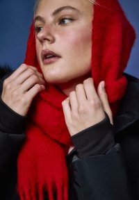 Rdeč pleteni šal s cofki, ovit okoli ramen, nošen s črno majico z dolgimi rokavi, poudarjajoč teksturo in žive barve.