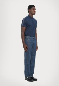 Hombre de pie mirando hacia la derecha, vestido con una camiseta polo azul marino con rayas estampadas en las mangas, jeans azules y zapatos de cuero negros, contra un fondo liso.