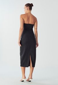 Žena stojící zády k fotoaparátu v černých midi šatech bez ramínek a bílých vysokých podpatcích na pozadí s jednobarevným světlým tónem.