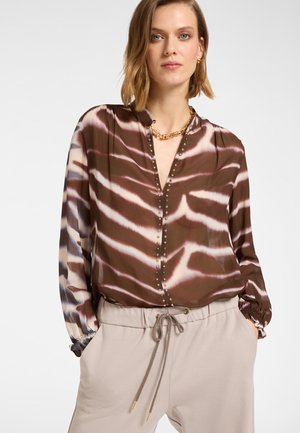 Blouse à motifs marron et crème avec un col en V, des boutons décoratifs et des poignets élastiques, associée à un pantalon beige clair avec cordon de serrage.