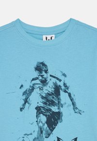 Ανοιχτό μπλε μπλουζάκι με γραφική εκτύπωση ενός ποδοσφαιριστή σε κίνηση, περιγραμμένου σε σκούρες μπλε αποχρώσεις, με κοντά μανίκια και στρογγυλή λαιμόκοψη.