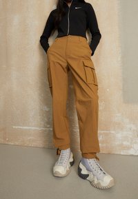 Bruna cargobyxor med flera fickor, matchade med kraftiga grå och beige sneakers. Svart kort zipjacka med logotypdetalj.