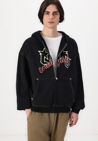 Svart hettegenser med glidelås, prydet med stjerner og tekstgrafikk i kremfarge og rødt. Den har en avslappet passform, kengurulommer og hvite sømdetaljer.