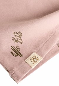 Helles rosa Stoff mit kleinen grünen Kaktusmotiven, an der Ecke mit einem strukturierten weißen Etikett, das ein Sonnendesign zeigt.