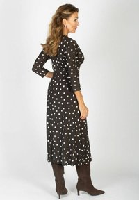 Kobieta z długimi falującymi włosami, ubrana w czarną sukienkę midi w białe kropki i brązowe botki na obcasie, stojąca w profilu z jedną ręką na biodrze.