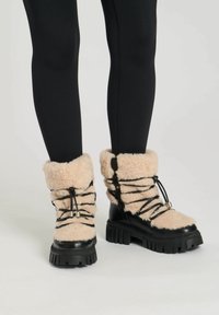 Botas negras forradas con piel de oveja, con suelas de goma gruesas, que presentan cordones y acentos negros, y un suave puño beige alrededor del tobillo.