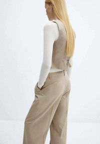 Beige taillierter Weste und passende weit geschnittene Hosen. Die Weste hat eine Rückenbindung und ärmelloses Design über einem weißen Langarmoberteil.
