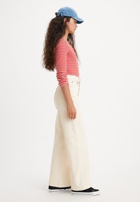 Jean crème taille haute et large avec une texture lisse. Assorti à un haut à manches longues rayé rouge et une casquette bleue, complété par des baskets noires.
