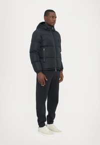 Black puffer jacket with a hood, featuring a front zipper and two side pockets, worn with black sweatpants and light sneakers.