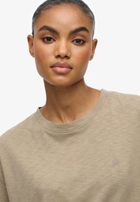 Beige t-shirt med rund hals i en blød tekstur, med et lille logo på brystet og et minimalistisk design.