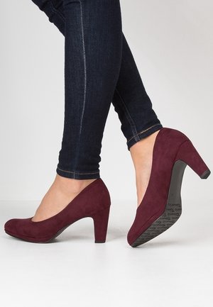 Burgundowe zamszowe szpilki z okrągłym noskiem, o teksturowanej powierzchni i umiarkowanej szpilce, noszone z ciemnymi dżinsami.