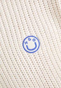 Crème-kleurige gebreide stof met een blauwe ronde patch met de tekst "HGO" en een gestileerde "U" in het midden.