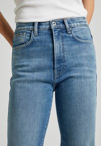 Detailní záběr na modré džíny střední výšky, které jsou nošeny s bílým žebrovaným topem na ramínka, zobrazuje detaily předních kapes a zapínání na knoflík.