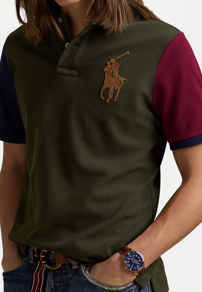 Polo shirt in donkergroen met kastanjebruine en marineblauwe mouwen, met een bruin geborduurd logo van een polo speler. Gemaakt met blauwe jeans en een ronde horloge.