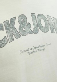 T-shirt chiaro con "CK & JON" in glitter argento e testo sottostante che recita "Creato a Copenaghen dal millenovecentonovanta."