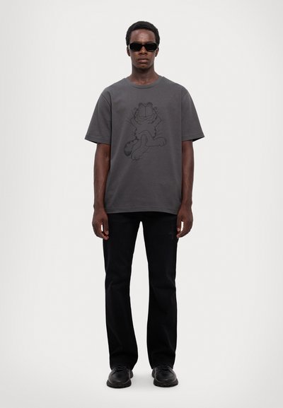 T-shirt en coton gris avec un graphique de dessin animé noir d'un chat, associé à un jean noir et des chaussures noires. Le mannequin porte des lunettes de soleil.
