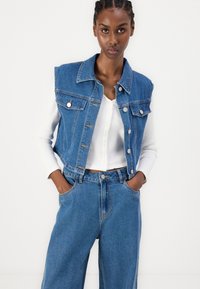 Denim brezrokavnik s klasičnim ovratnikom, gumbnim zapiranjem in dvema žepoma na prsih, nošen preko rebraste bele majice in v kombinaciji z širokimi kavbojkami.