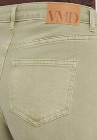 Κοντινό πλάνο πίσω τσέπης από φως πράσινο τζιν με γαζωμένα άκρα και καφέ δερμάτινη ετικέτα με λογότυπο VMD στη μέση του παντελονιού.